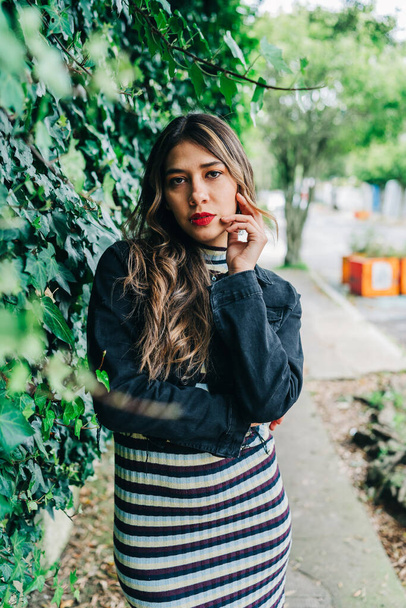 in the city street a young latin woman with long blonde hair, wears makeup with red lips and fashionable clothes, beauty with nature by her side, lifestyle - Photo, image