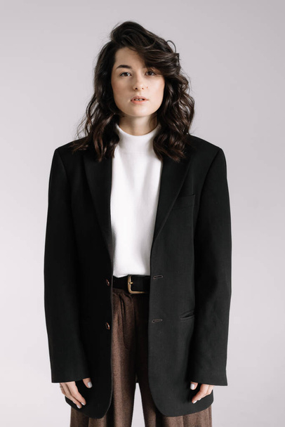 Fashion studio photo of stylish European brunette woman in in long baggy pants and a black jacket posing on white background. Trendy office style in clothes.  - Photo, Image
