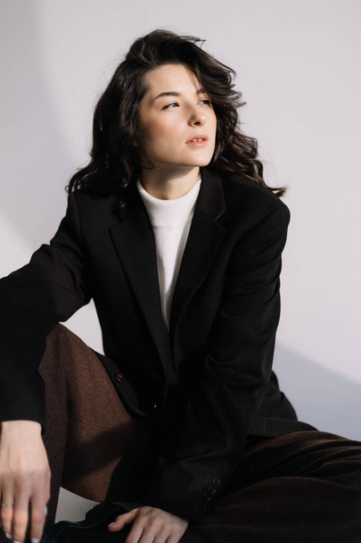 Fashion studio photo of stylish European brunette woman in in long baggy pants and a black jacket posing on white background. Trendy office style in clothes.  - Zdjęcie, obraz