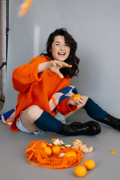 young girl sitting on the floor in a photo studio with tangerines in her hands, posing for a photo, throwing tangerines into the camera, laughing and having fun. Bright emotions - 写真・画像