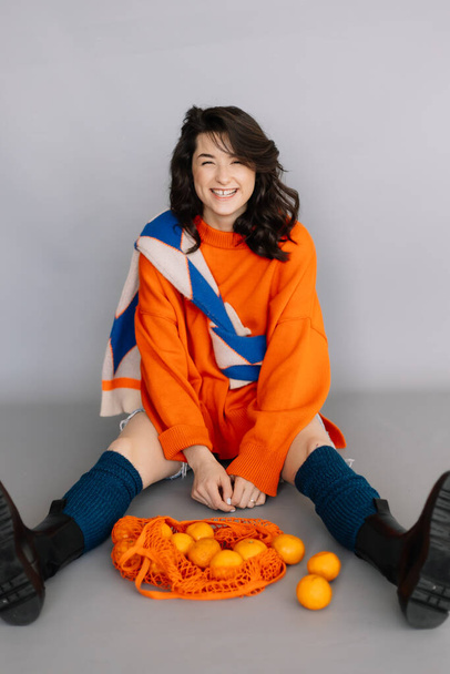 stylish woman dressed in orange knitted jacket posing for a photo in a photo studio on a gray paper background. Bright emotions, poses, modern concept - Фото, зображення
