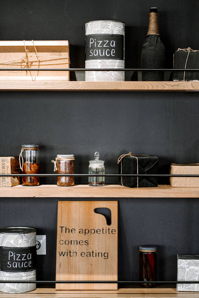 Стильная посуда хранится на деревянной полке на фоне черной стены. Фото объекта интерьера. Резка доски и банки специй на кухне - Фото, изображение