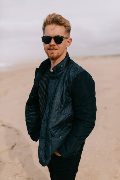 Stylish European young blond man in sunglasses is wearing dark blue jacket is posing at camera on the shore near the ocean on blue sky background . High quality photo - Foto, Imagem