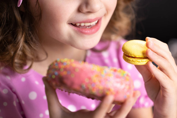 Caucasian child school girl eating donut and macaron at the same time. Adorable kid girl biting piece of donut macaron while sitting at table at home. Sweet addiction concept - Photo, Image