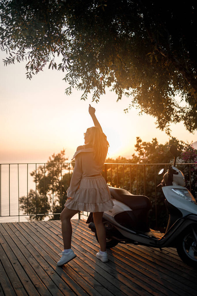 Una joven mujer hermosa con ropa de verano se encuentra en el fondo de una puesta de sol naranja cerca de la ciudad ciclomotor. enfoque selectivo - Foto, imagen