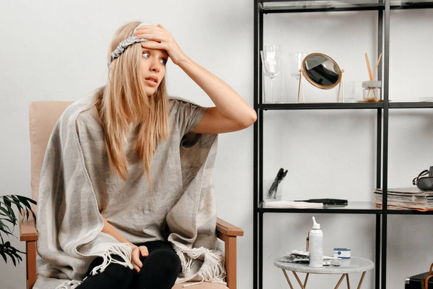 Blonde hair girl with a sleeping mask on the face is sitting in a chair. Young woman is holding a hand on the head because she has strong headache - Foto, Bild