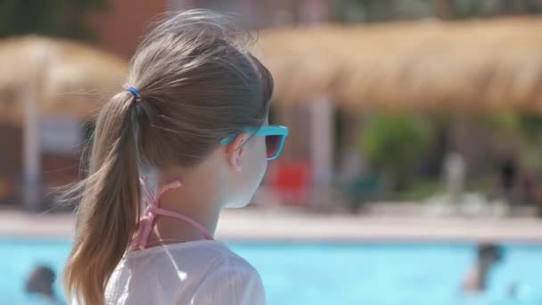Jovem menina alegre descansando no lado da piscina com água azul clara no dia ensolarado de verão. Conceito de férias tropicais - Filmagem, Vídeo