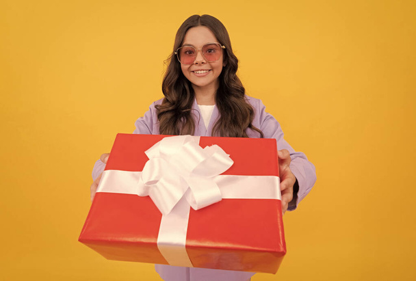trendy lächelndes Kind in Sonnenbrille und Anzug halten Geschenkbox auf gelbem Hintergrund, Geschenk - Foto, Bild
