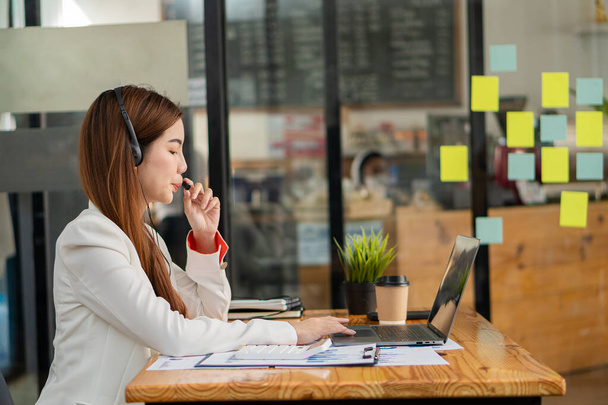 Asian woman wearing headphones using laptop talking to a coworker about sales report in video conferencing while working at a coffee shop. financial accounting business concept - Foto, immagini
