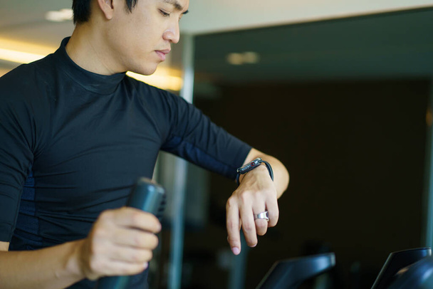 Happy sportsman using a smartwatch or exercise tracker on his wrist to track an exercise, sport man touching a smartwatch screen while exercising on a elliptical exercise machine. Serious workout. - Foto, imagen