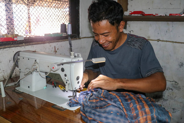 アジア人の男はミシンで服を縫う。ワークショップでミシンや布で働く中年男性の仕立屋 - 写真・画像