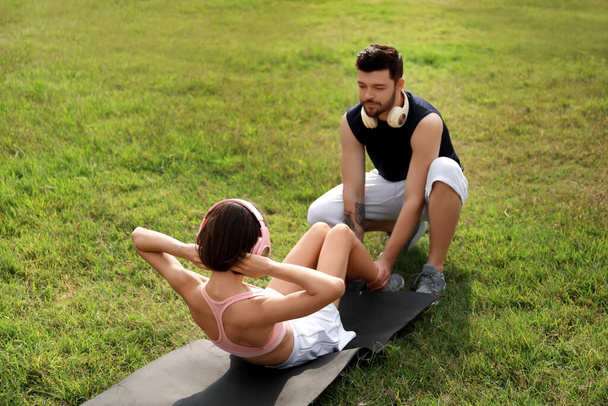 Спортивная молодая женщина в наушниках делает упражнения на прессе с мужем в парке - Фото, изображение