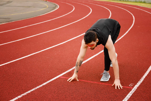 Αθλητικός νεαρός με γυαλιά ηλίου ετοιμάζεται να τρέξει στο στάδιο. - Φωτογραφία, εικόνα