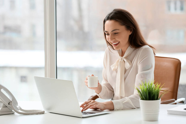 Jovem secretária bebendo café enquanto trabalhava no laptop no escritório - Foto, Imagem