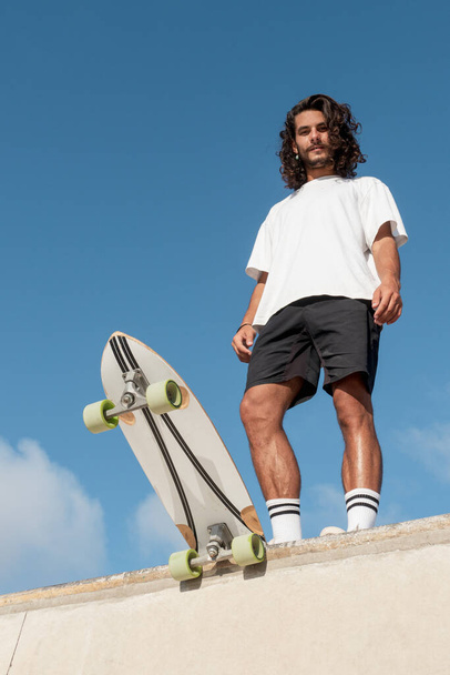 Young handsome caucasian skater at the top of the skate ramp. He has long black hair and a beard and he is wearing summer clothes - Photo, Image