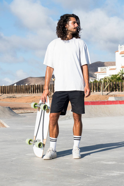 Young male caucasian skater standing at the skate park. He is holding his surfskate skateboard and whearing summer clothes. He has long black hair. - Photo, Image