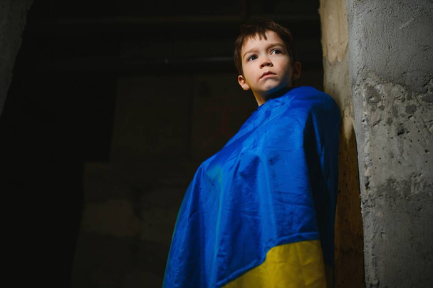 A sad Ukrainian boy wrapped in a Ukrainian flag, hiding from bombings in a bomb shelter in the city of Mariupol or Kiev. Russia's aggression. The war in Ukraine and 2022. - Foto, Bild
