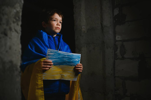A sad Ukrainian boy wrapped in a Ukrainian flag, hiding from bombings in a bomb shelter in the city of Mariupol or Kiev. Russia's aggression. The war in Ukraine and 2022. - Valokuva, kuva