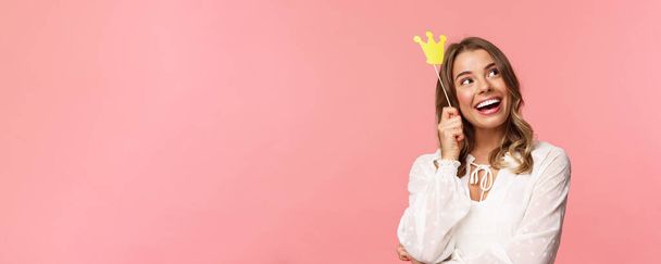 Spring, happiness and celebration concept. Close-up portrait of dreamy beautiful young blond girl imaging something cute and romantic, smiling look up daydreaming with crown on stick, pink background - Foto, immagini
