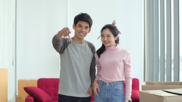 Mladý ženatý asijský pár s krabicemi a drží ploché klíče a dívat se na fotoaparát v novém domově. Stěhování domu. - Záběry, video