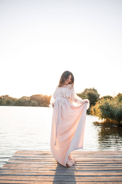 Una joven embarazada con un vestido de gasa se encuentra en un muelle junto al río en el contexto de una puesta de sol naranja - Foto, imagen
