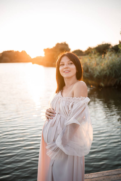 Eine junge schwangere Frau im Chiffonkleid steht auf einem Steg am Fluss vor der Kulisse eines orangefarbenen Sonnenuntergangs - Foto, Bild