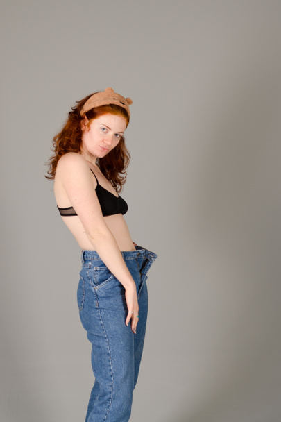 Portrait of beautiful young woman in black sports top and jeans with red curly hair, freckles and blue eyes against gray background. Lifestyle. Beauty.  - Φωτογραφία, εικόνα