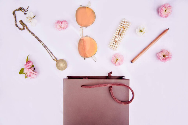 Splash γυναικείων αξεσουάρ από καφέ τσάντα με ροζ λουλούδια. Σακούλα δώρου. Γυαλιά ηλίου, αξεσουάρ, φουρκέτα, σκουλαρίκια, μενταγιόν λαιμού, μολύβι μακιγιάζ σε λευκό φόντο. 8 Μαρτίου έννοια. - Φωτογραφία, εικόνα