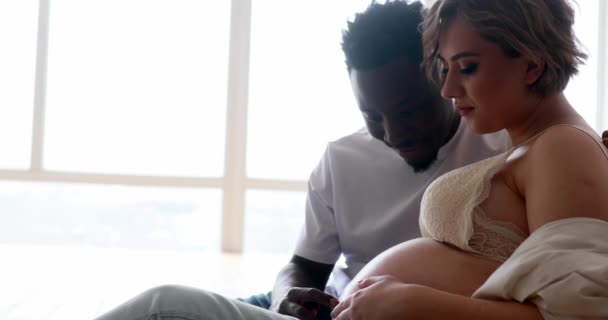 A fiatal, boldog afrikai férfi és a kaukázusi terhes nője együtt ülnek, jól érzik magukat és boldogan mosolyognak. Fajok közötti házasság. Anyaság és terhesség. - Felvétel, videó