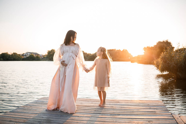 A pregnant woman with her daughter against the background of water at sunset, a woman with a belly enjoys the sunset by the river - Photo, Image
