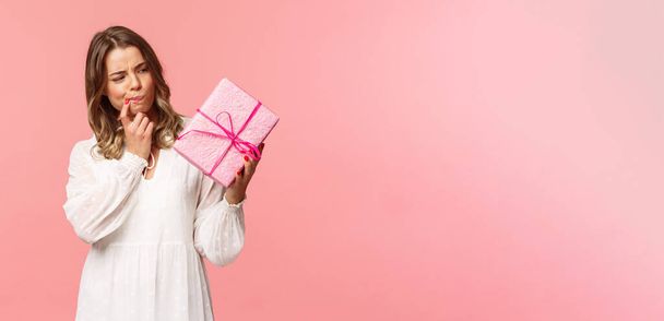 Holidays, celebration and women concept. Portrait of thoughtful curious blond girl thinking whats inside gift box, look pondering say hmm as making assumpion, standing pink background - Foto, afbeelding