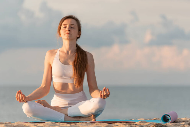 Молодая красивая женщина в позе лотоса медитирует на пляже. Женщина практикует йогу на морском фоне - Фото, изображение