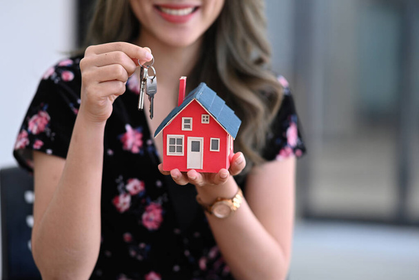 Smiling asian woman holding house model and house key in hand. Real estate investment and insurance concept. - Foto, imagen