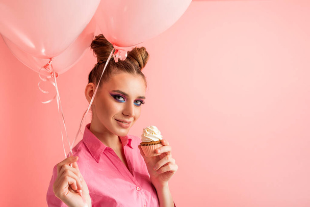 Girl with professional pink makeup and balloons enjoys delicious cupcakes. Portrait of woman with sweets on pink background - Fotó, kép