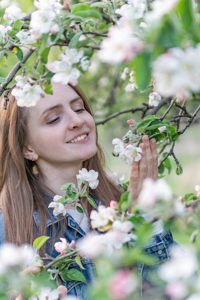 Uśmiechnięta piękna kobieta w pobliżu kwitnącej jabłoni cieszy się aromatem kwiatów. Portret dziewczynki w wiosennym ogrodzie. Rama pionowa - Zdjęcie, obraz