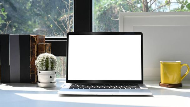 Ordenador portátil Mockup, marco de imagen, libros y cactus en la mesa blanca en la oficina del hogar brillante. - Foto, Imagen