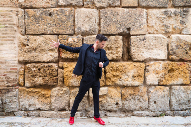 Ένας τσιγγάνος που χορεύει φλαμένκο, νέος και όμορφος ντυμένος με μαύρα και κόκκινα παπούτσια ποζάρει και χορεύει σε ένα φόντο πέτρινου τοίχου. Φλαμένκο πολιτιστική κληρονομιά της ανθρωπότητας. - Φωτογραφία, εικόνα