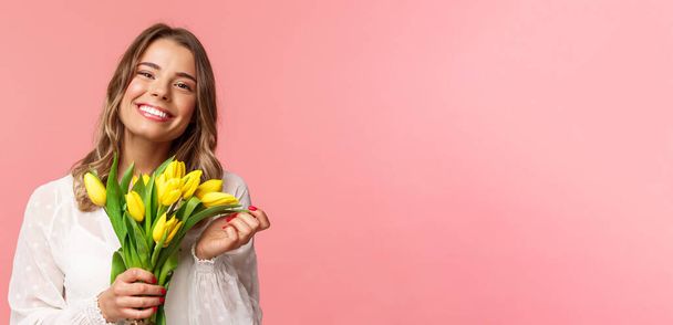 Spring, happiness and celebration concept. Close-up of lovely happy young blond girl in white dress, daydreaming after perfect date, holding yellow tulips and smiling at camera, pink background - Photo, Image