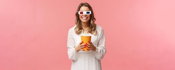 Leisure, going-out and spring concept. Portrait of happy carefree blond girl in white dress enjoying watching movie in 3d, wearing glasses eating popcorn and smiling, attend cinema, pink background - Fotó, kép