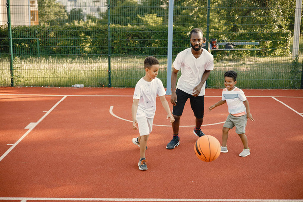 Чёрный отец с двумя сыновьями, играющими в баскетбол на баскетбольной площадке. - Фото, изображение