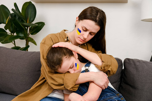 Close up of caring young mother cuddling sad little daughter with flag on face sitting together on grey couch at home. Peace, no war, help, stop russian aggression. Invasion of Russia in Ukraine - Foto, Imagen