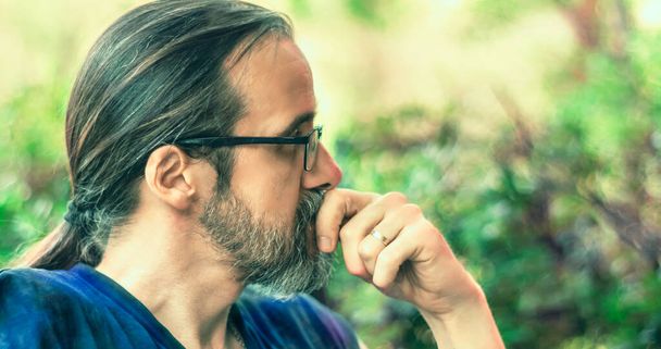 Retrato de um homem adulto médio com barba, óculos e cabelos longos em um rabo de cavalo, olhando pensativo. Vista lateral. Foco seletivo - Foto, Imagem