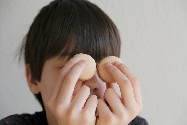 close-up of part of the child's face, boy 10-12 years old Asian-European appearance, makes binoculars from macaroons, concept of vision examination, childhood, sweet life, emotional development - Photo, image