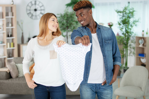 cheerful couple holding baby clothing at home - Photo, Image