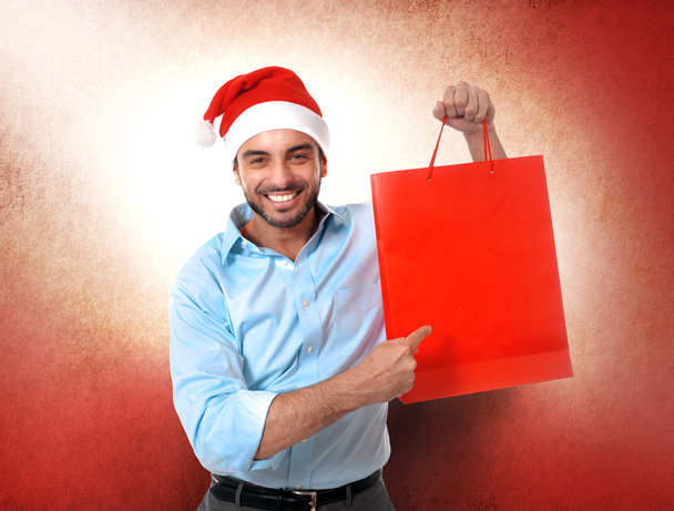 happy young handsome man wearing santa hat holding red shopping  - Фото, зображення