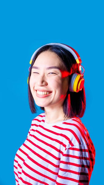 音楽を聴く青い背景に孤立した穏やかなアジアの女性ワイヤレスヘッドフォンの笑顔-垂直バナー - 写真・画像