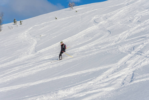 Donna con i capelli castani, con indosso abbigliamento da sci e bagaglio sciare giù per una bella collina di neve con un sacco di tracce sulla neve, vista dal lato da lontano, Stryn Norway, orizzontale - Foto, immagini