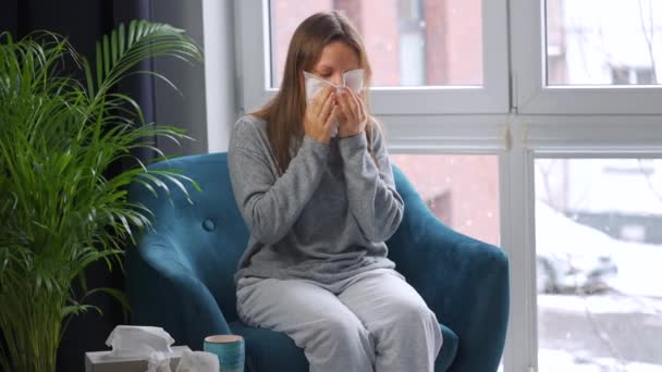 Mulher insalubre senta-se em uma cadeira e espirra ou sopra o nariz em um guardanapo porque ela tem um resfriado, gripe, coronavírus. Está nevando lá fora. - Filmagem, Vídeo