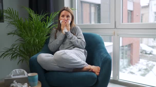 Az egészségtelen nő ül egy székben, és tüsszent vagy fújja az orrát egy szalvétába, mert megfázott, influenzás, koronavírusos. Havazik odakint. - Felvétel, videó