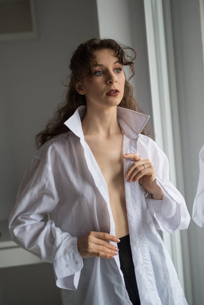 Attractive sexy brunette in white shirt posing provocatively, near a window,studio shot. Portrait of sensual woman with long hair, in classic boudoir scene , looked out the window - Photo, Image